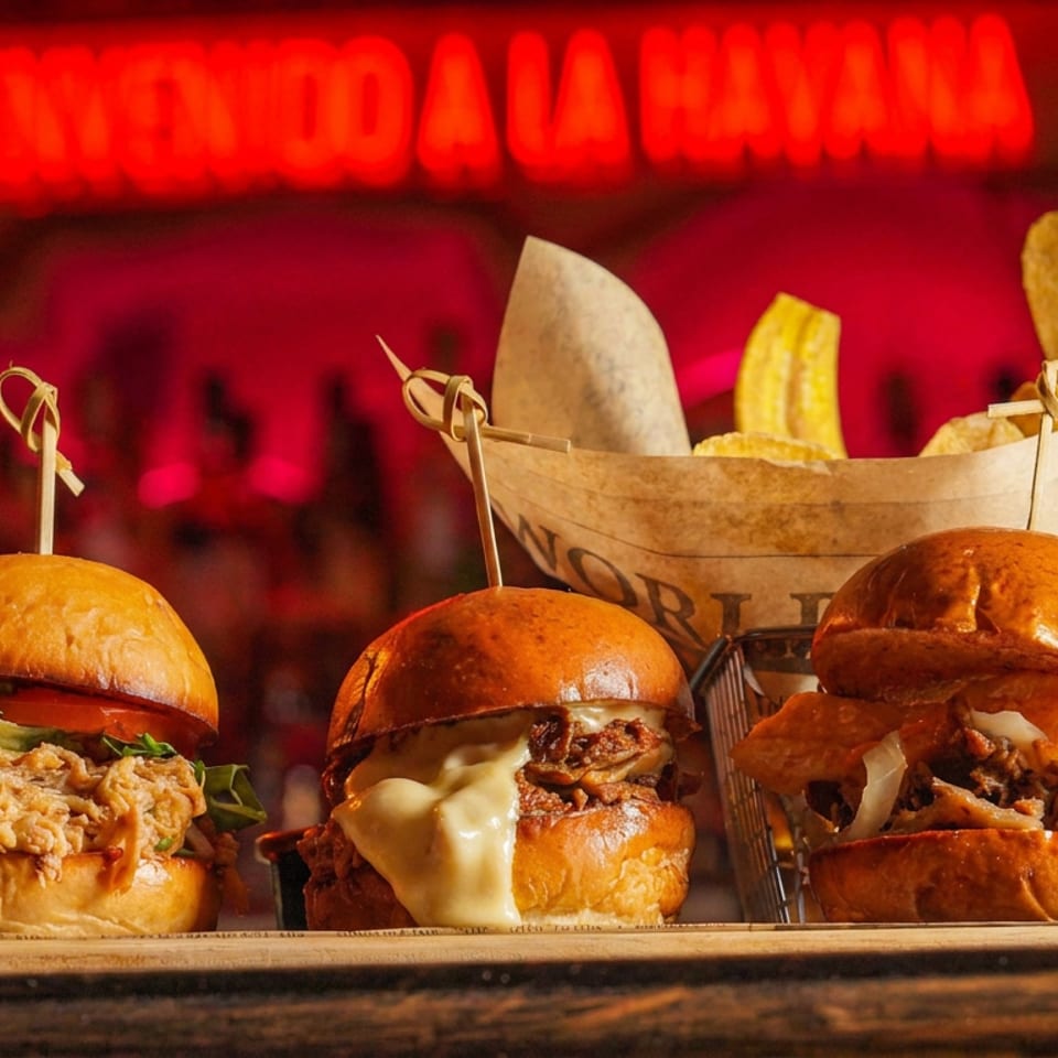 Three gourmet burgers with crispy toppings sit on a wooden board beside a basket of fries in a warmly lit bar.