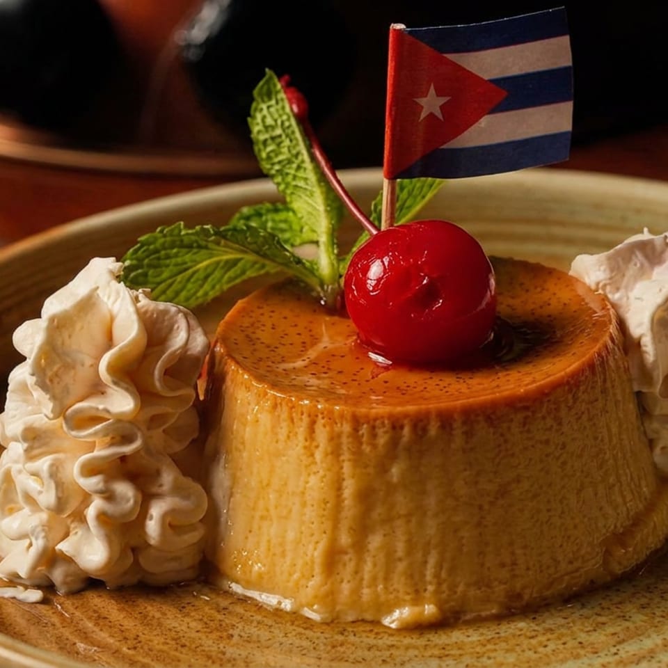 Caramel flan topped with a cherry and Cuban flag, served with whipped cream and mint on a ceramic plate.