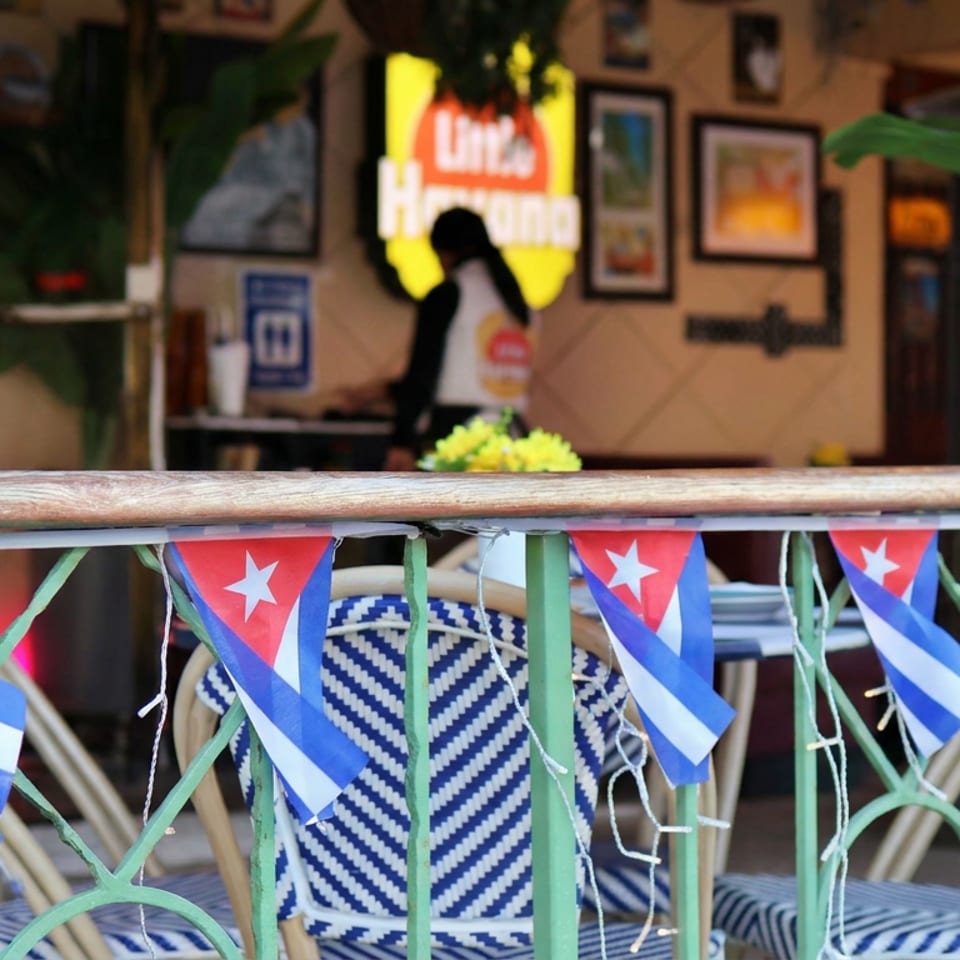 Cuban flags decorate a green patio railing outside a tropical restaurant with blue-and-white chairs, plants, framed artwork, and a blurred person.