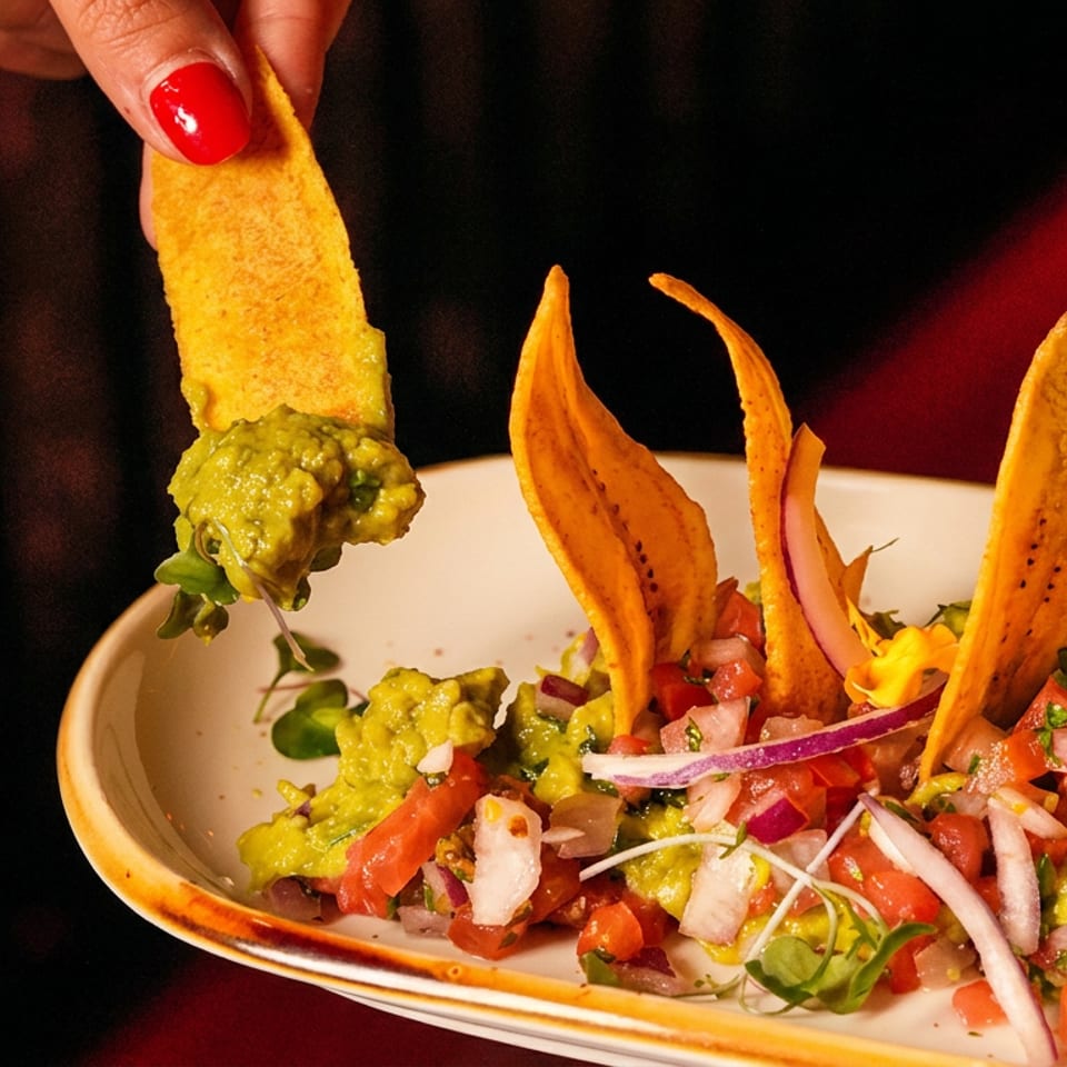 Tortilla chips with guacamole, pico de gallo, onions, and microgreens served on a plate, with one chip held above.