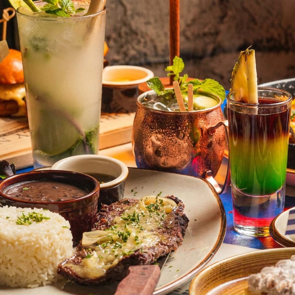 A restaurant table displays grilled steak with rice, tacos, salad, sauces, and colorful cocktails.