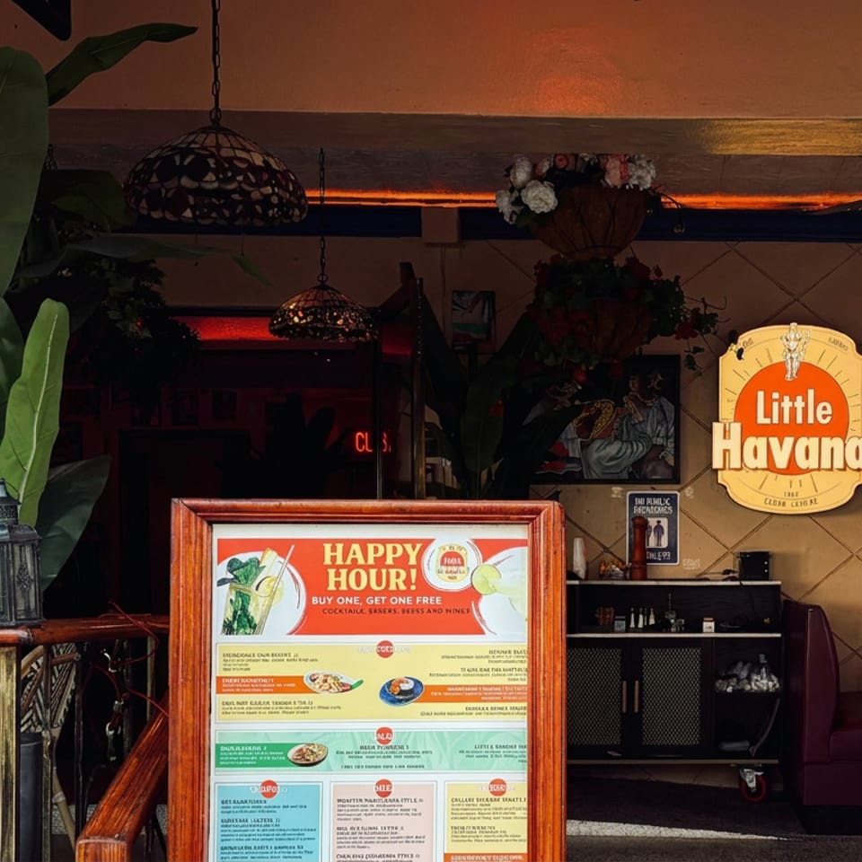 Dimly lit Little Havana restaurant interior with tropical plants, hanging lamps, framed artwork, and a prominently displayed happy hour menu.