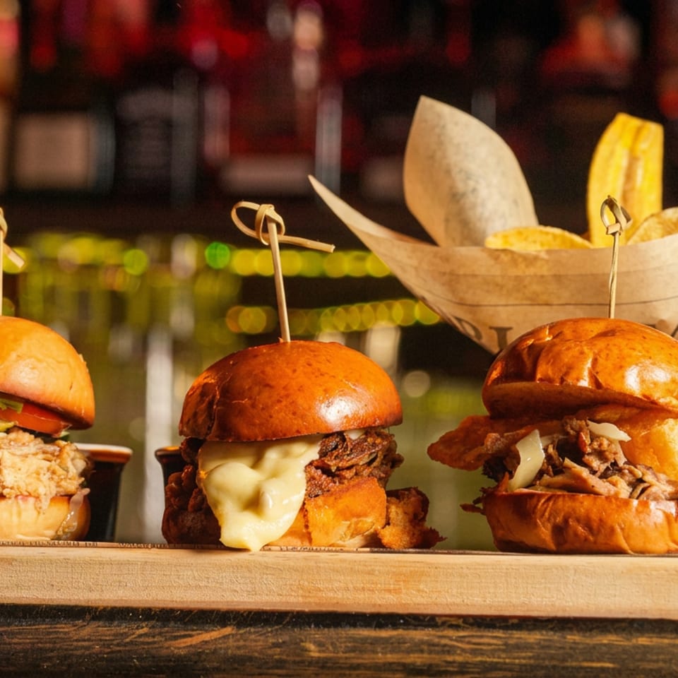 Three gourmet burgers on brioche buns sit on a wooden board beside a basket of golden fries in a warmly lit bar.