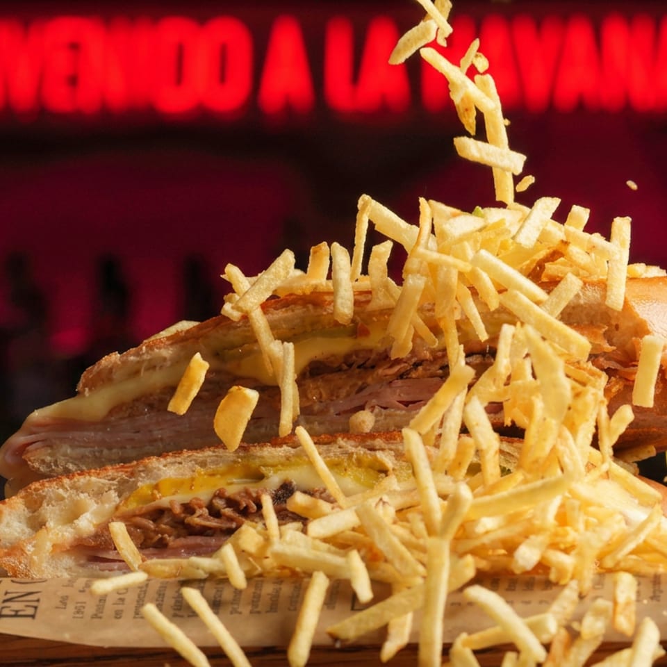 Stacked meat sandwiches topped with crispy shoestring fries, displayed on newspaper against a dim restaurant backdrop with red neon signage.
