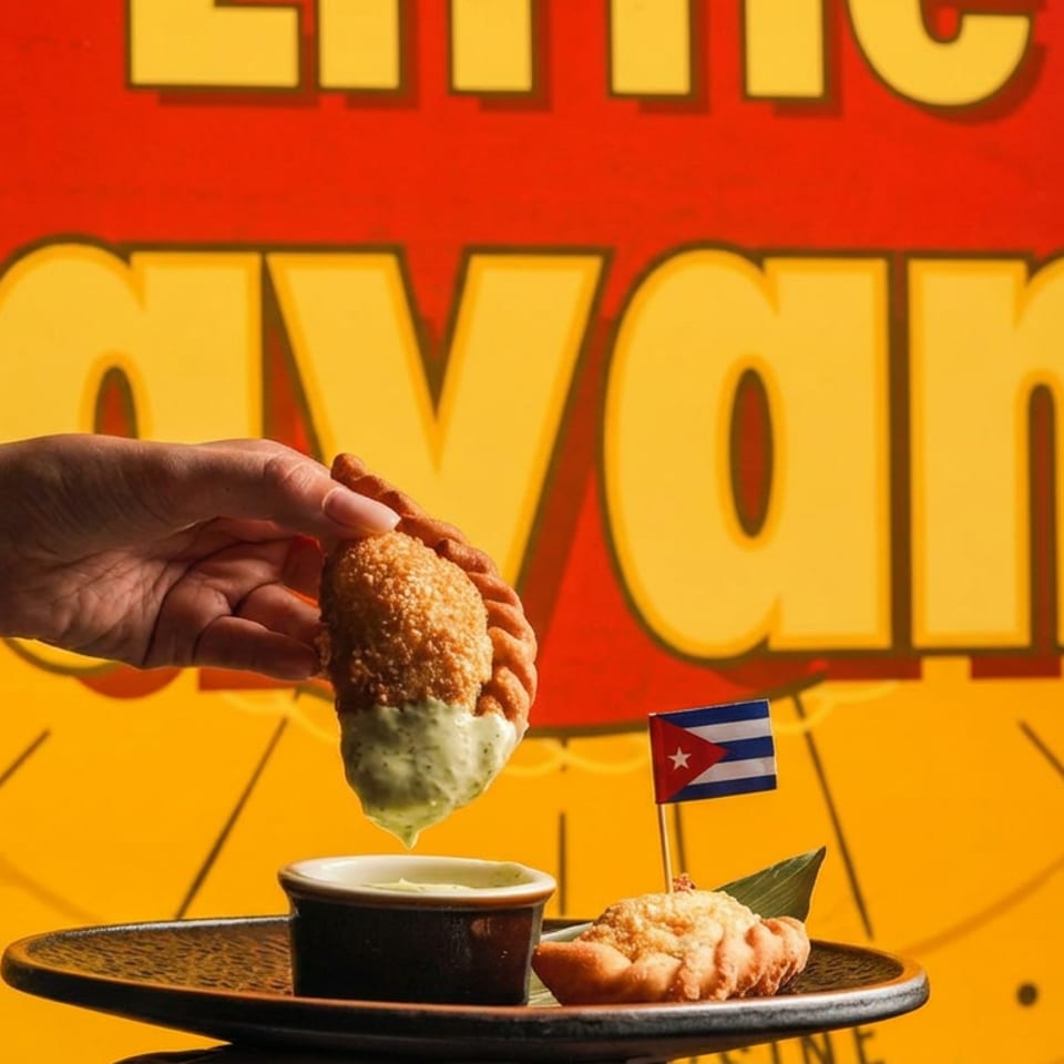 A hand dips a breaded appetizer into sauce above a plate, with a Cuban flag and colorful Havana-themed backdrop.