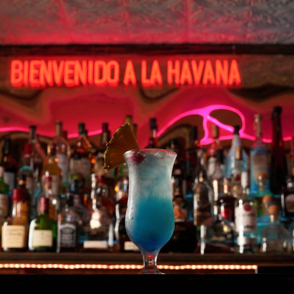 Blue cocktail garnished with a dried citrus slice sits before a liquor-filled bar beneath a glowing “Bienvenido a La Havana” sign.