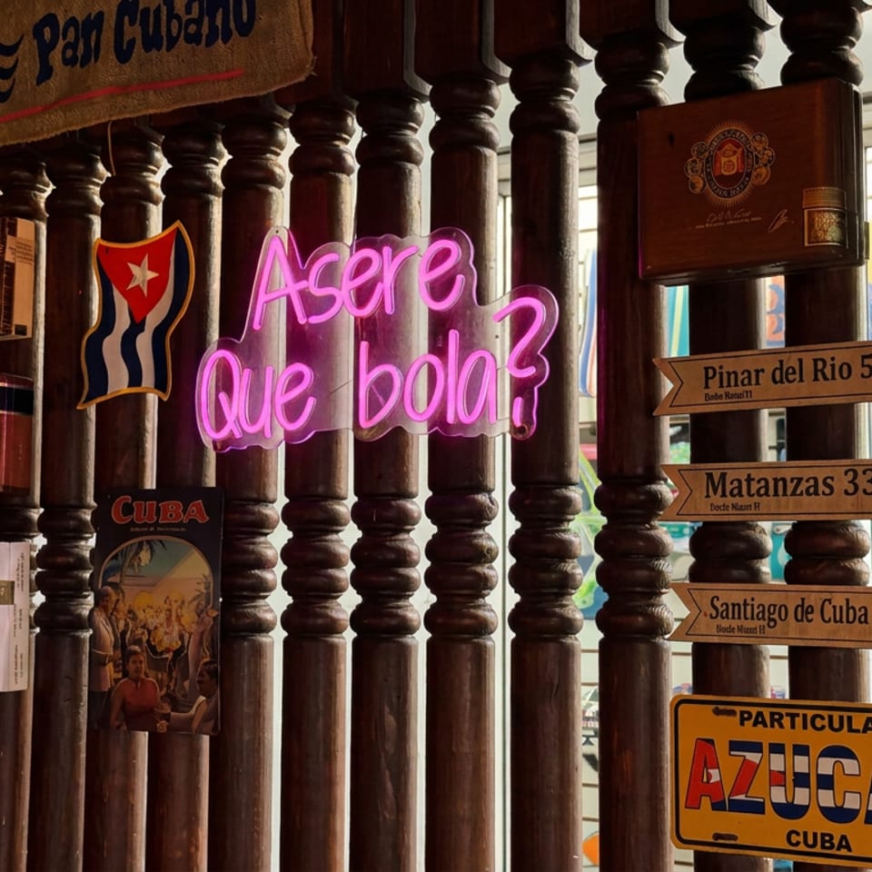 Cuban-themed restaurant display with neon “Asere Que Bola?” sign, cigar boxes, flags, and directional signs to Cuban cities.